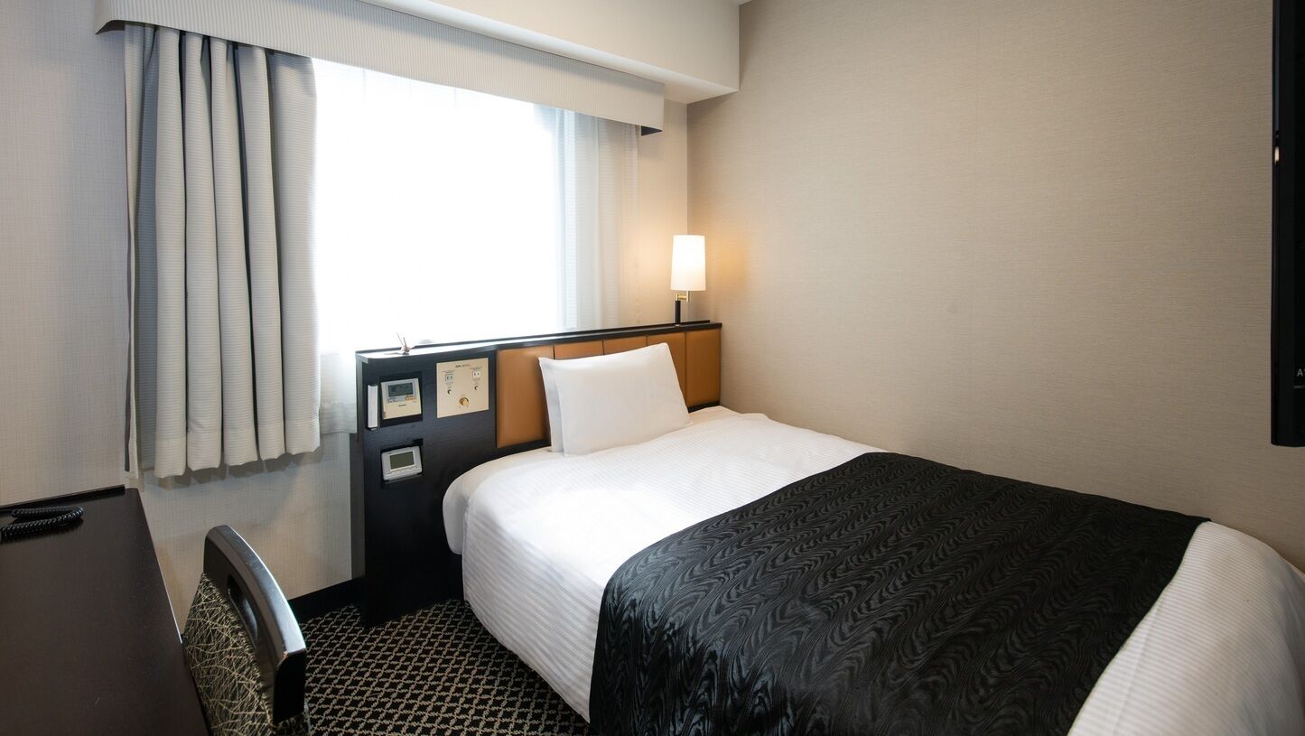 Een hotelkamer met een bed, bureau, stoel, raam met gordijnen en een wandlamp.