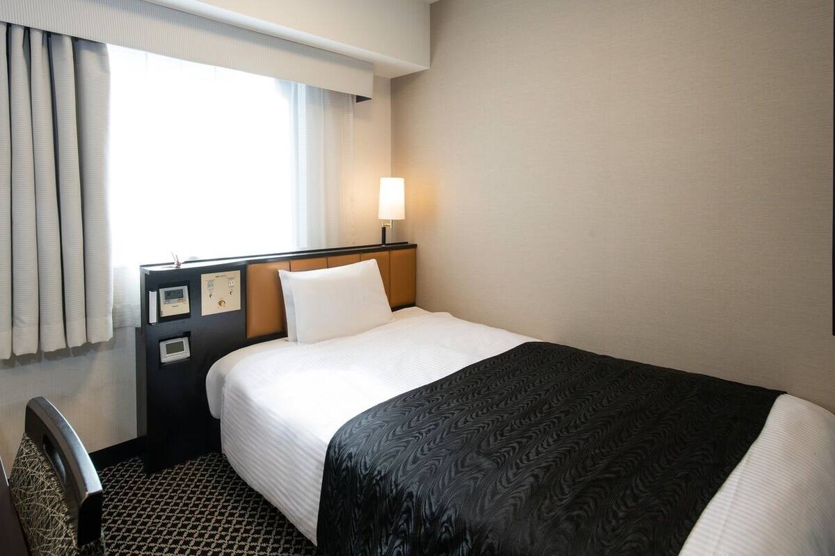 A hotel room with a bed, a desk, a chair, a window with curtains, and a wall-mounted lamp.