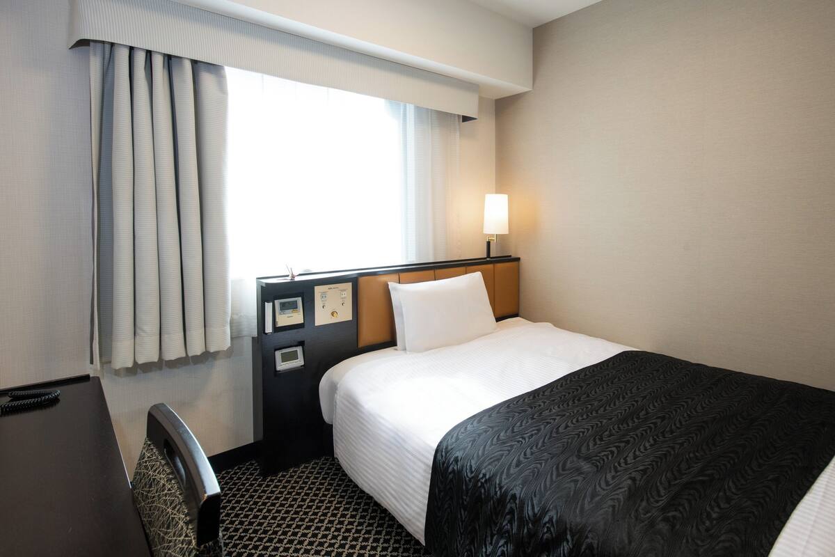 A hotel room with a bed, a desk, a chair, a window with curtains, and a bedside table with a lamp.