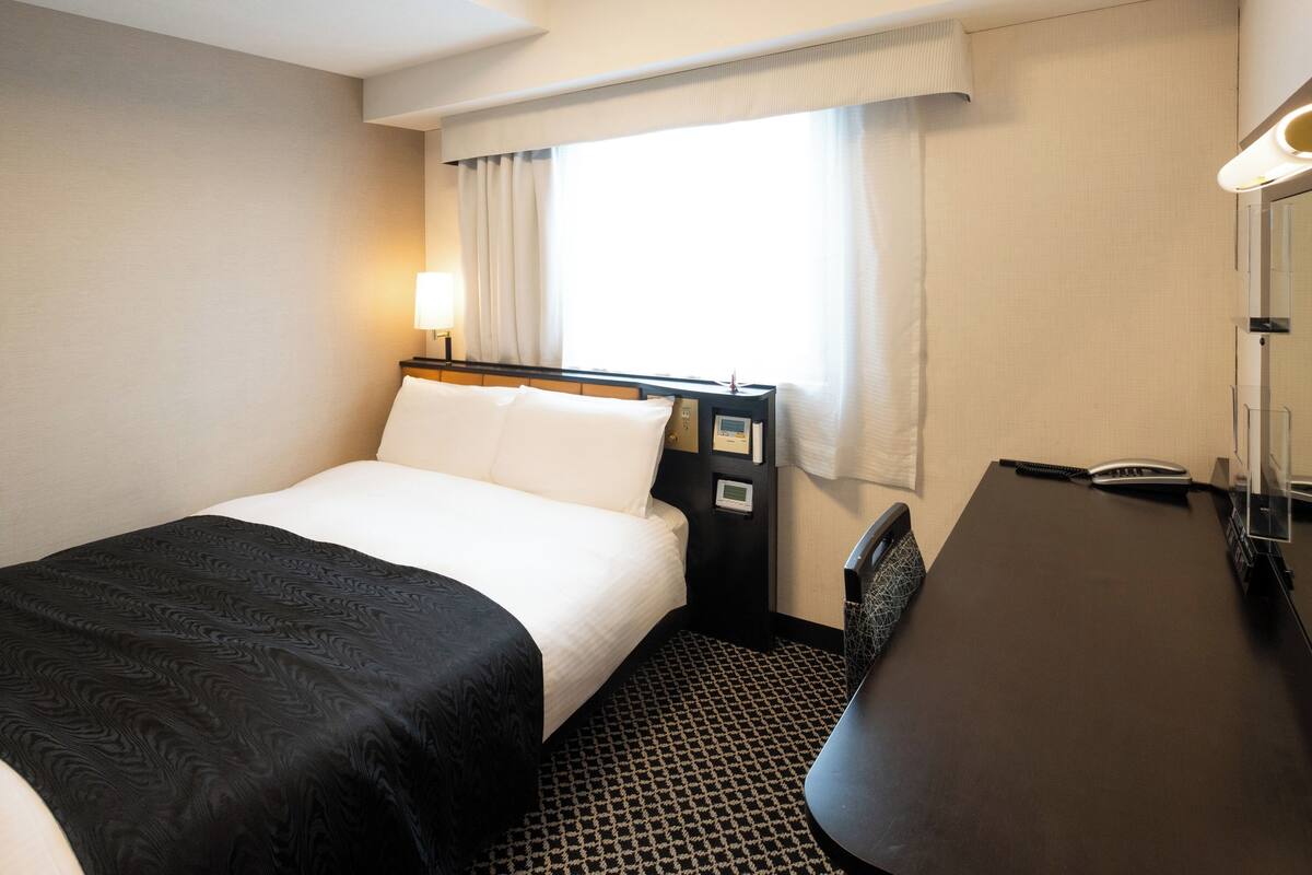 A hotel room with a bed, bedside table, lamp, and a desk with a chair.