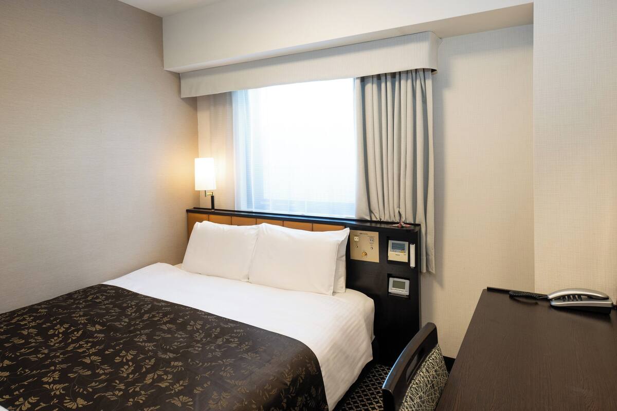 A hotel room with a bed, a desk, a lamp, a phone, and a window with curtains.