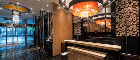 A hotel lobby with a reception desk, a chandelier, and a glass elevator entrance.