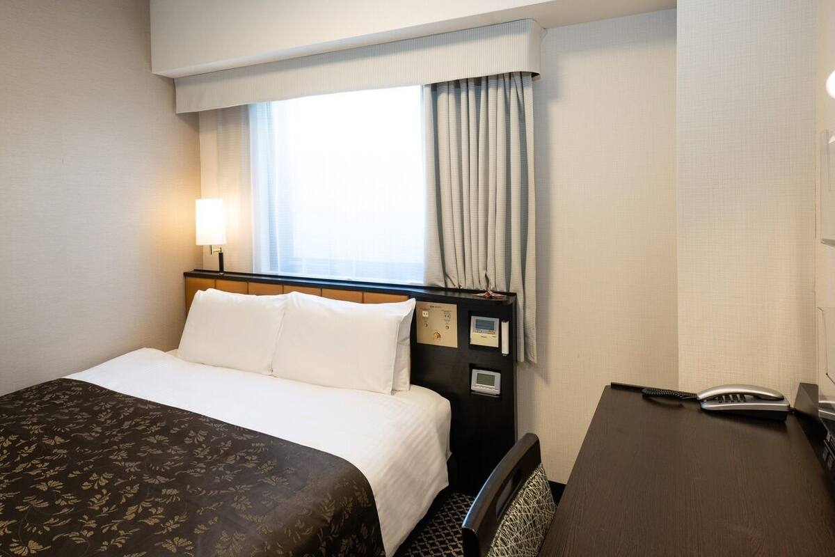 A hotel room with a bed, bedside table, lamp, and window with curtains.