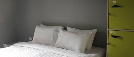 A bed with white bedding and pillows, a grey wall, and two green storage units with numbers 3701 and 3703.