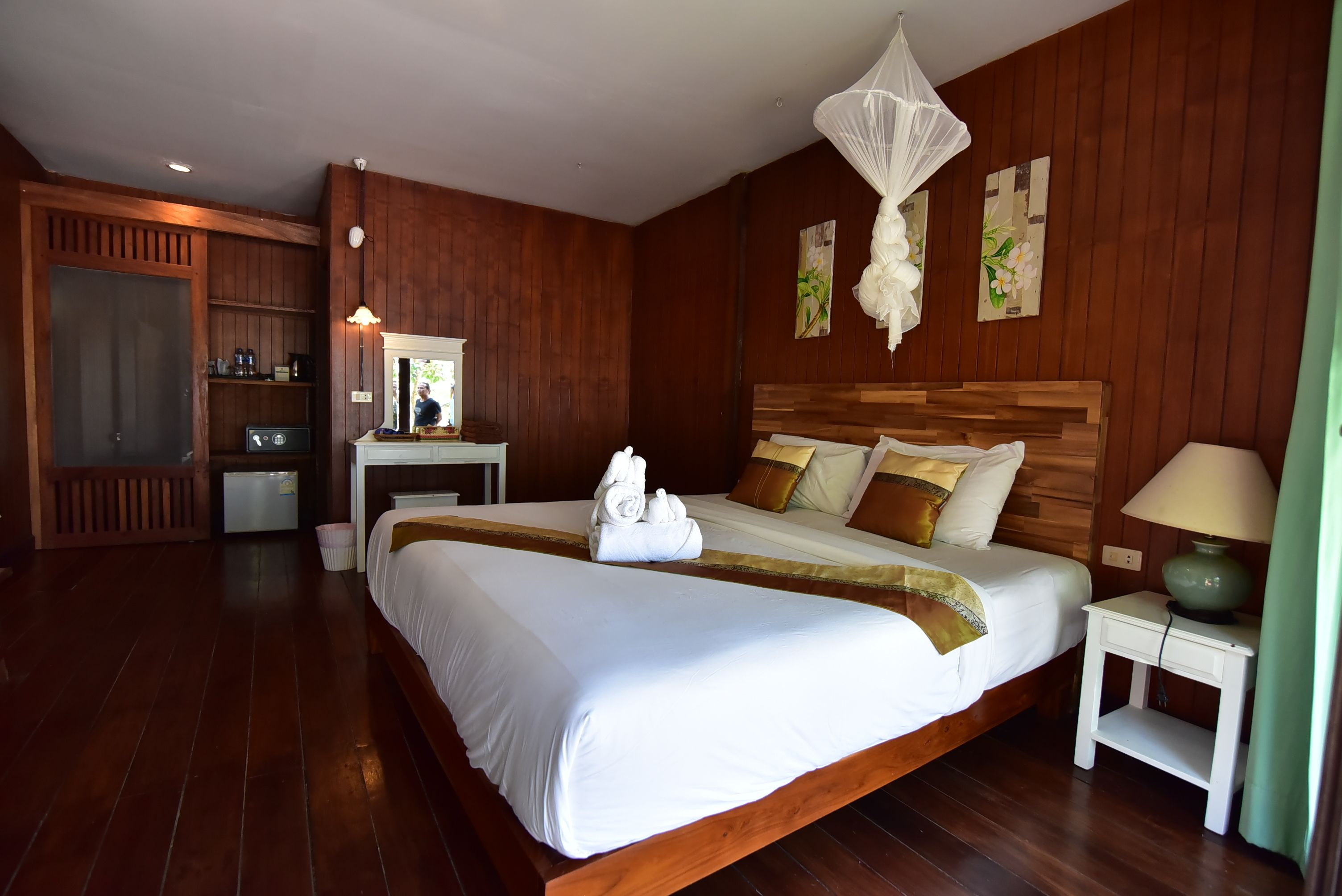 A hotel room with a large bed, wooden walls, a bedside table with a lamp, and a small desk with a mirror and coffee machine.