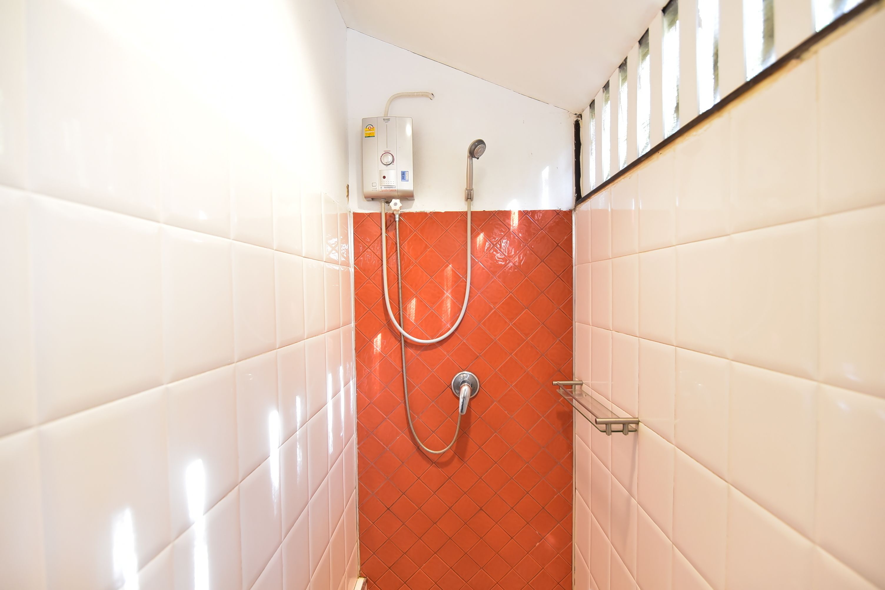 Un baño con paredes cubiertas de azulejos blancos y rojos, una ducha con cabezal de ducha manual y un calentador de pared.