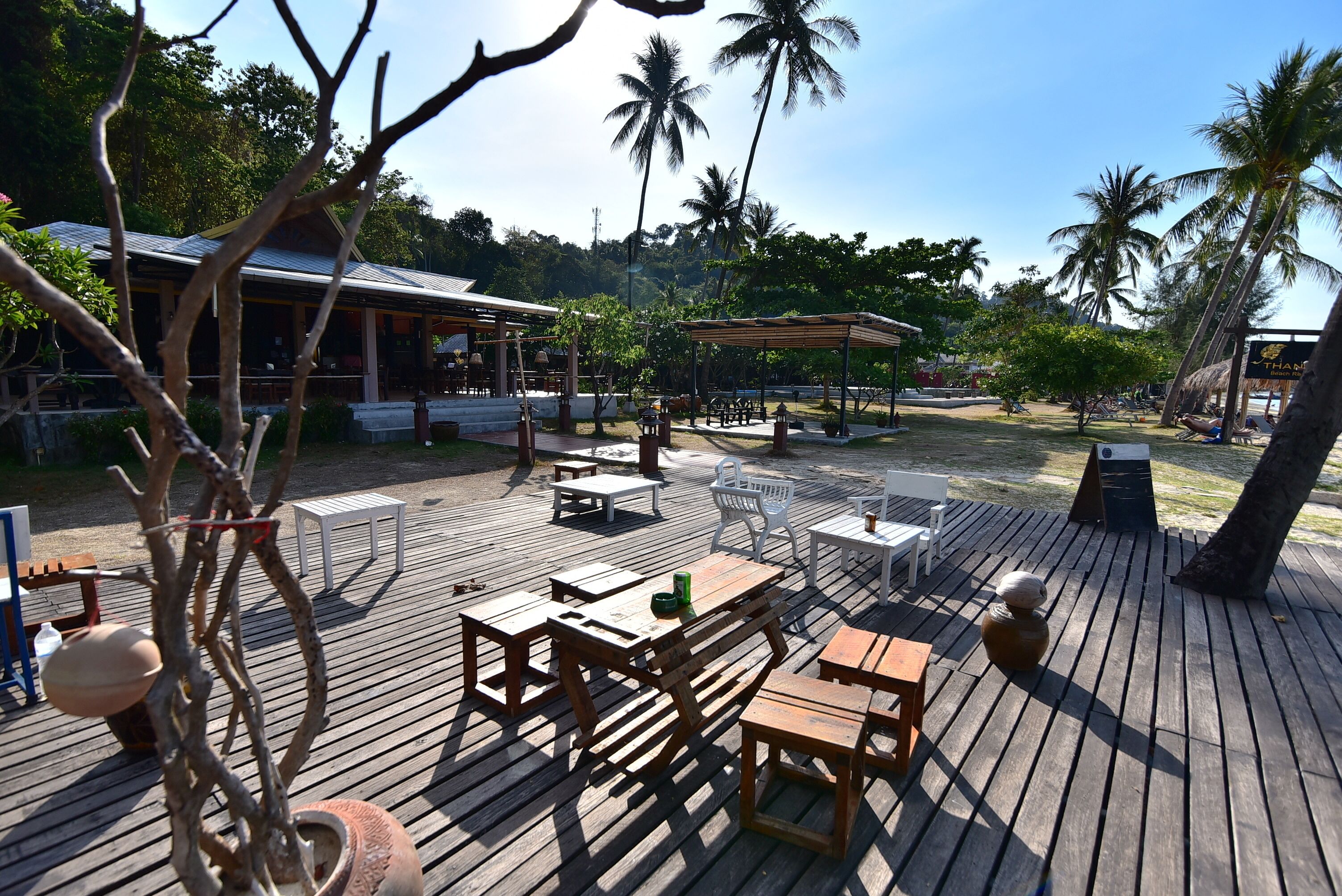 A wooden deck with outdoor seating, including tables and chairs, surrounded by palm trees and a building with a thatched roof.