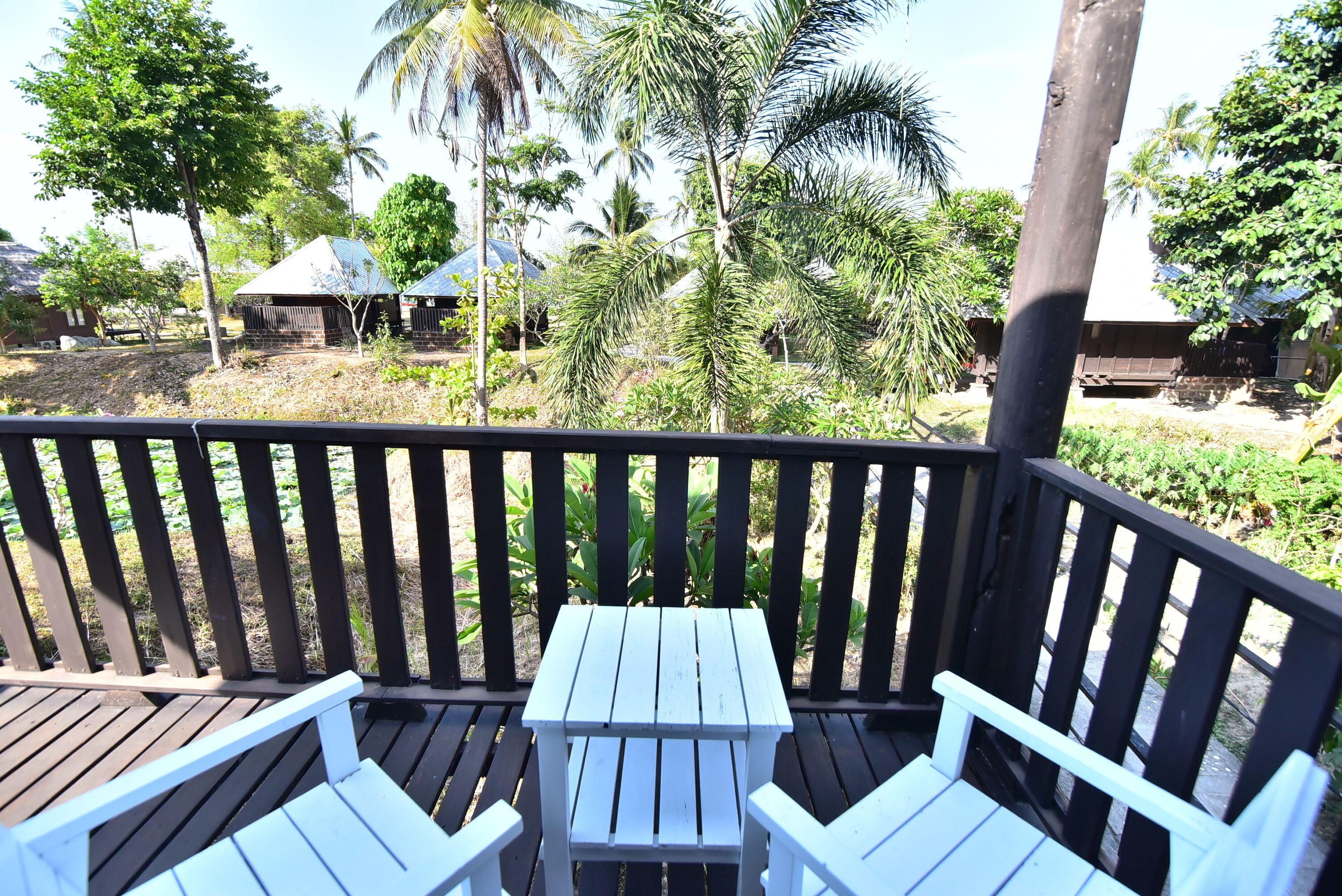 Una terraza de madera con una mesa y dos sillas, con vistas a un paisaje tropical con palmeras y un pequeño edificio.