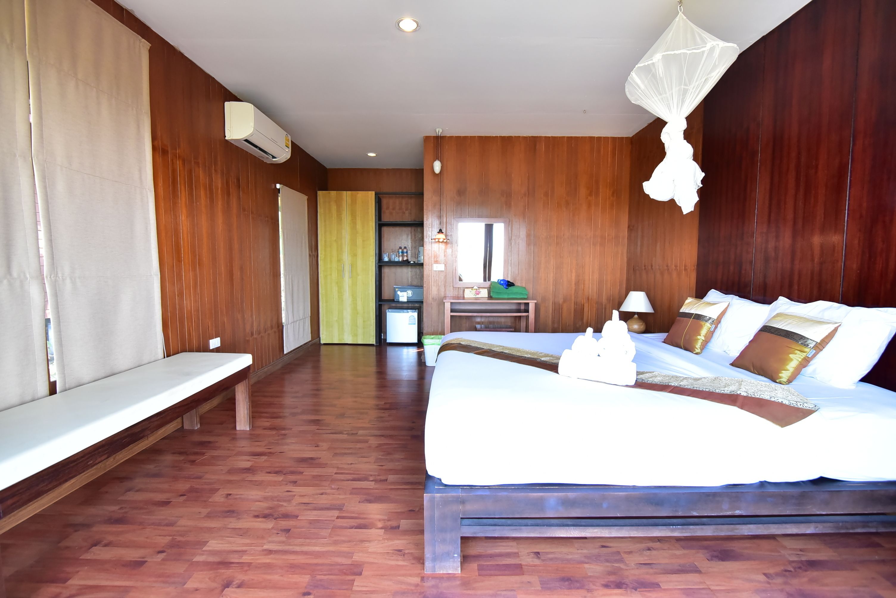 A hotel room with a wooden floor, a bed with white bedding, a bench, a desk with a chair, and a window.