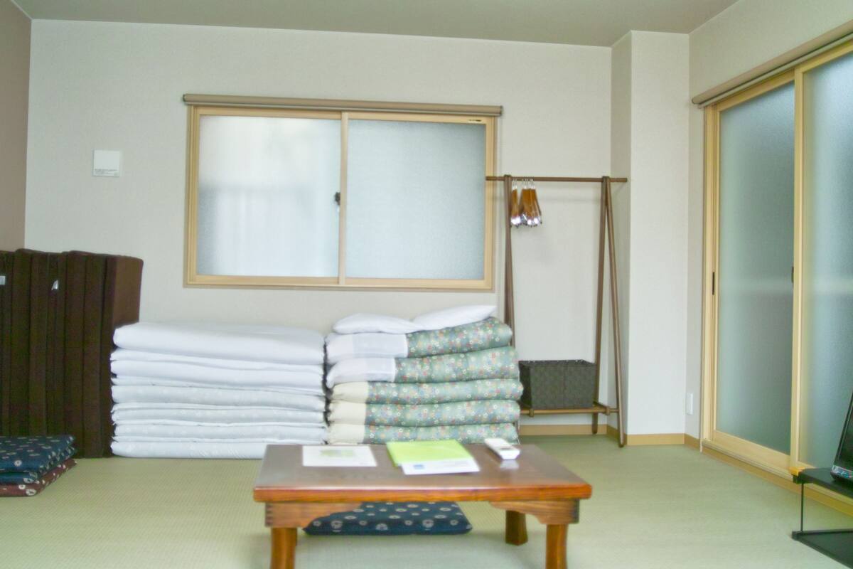 A room with a large window, a table with papers, a stack of folded blankets, and a futon bed.