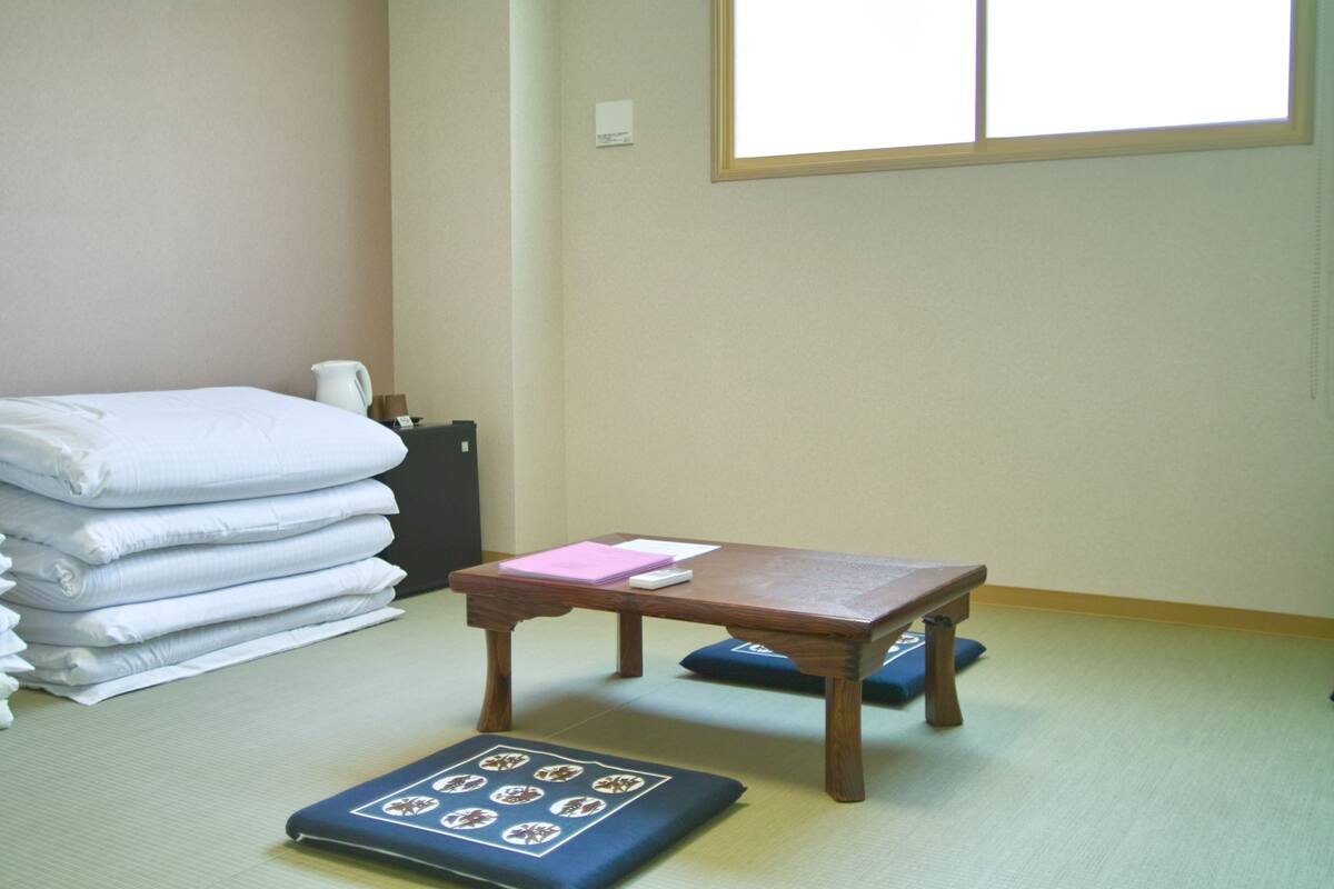 A room with tatami mats, a wooden table with a cushion, a stack of folded towels, and a window with blinds.