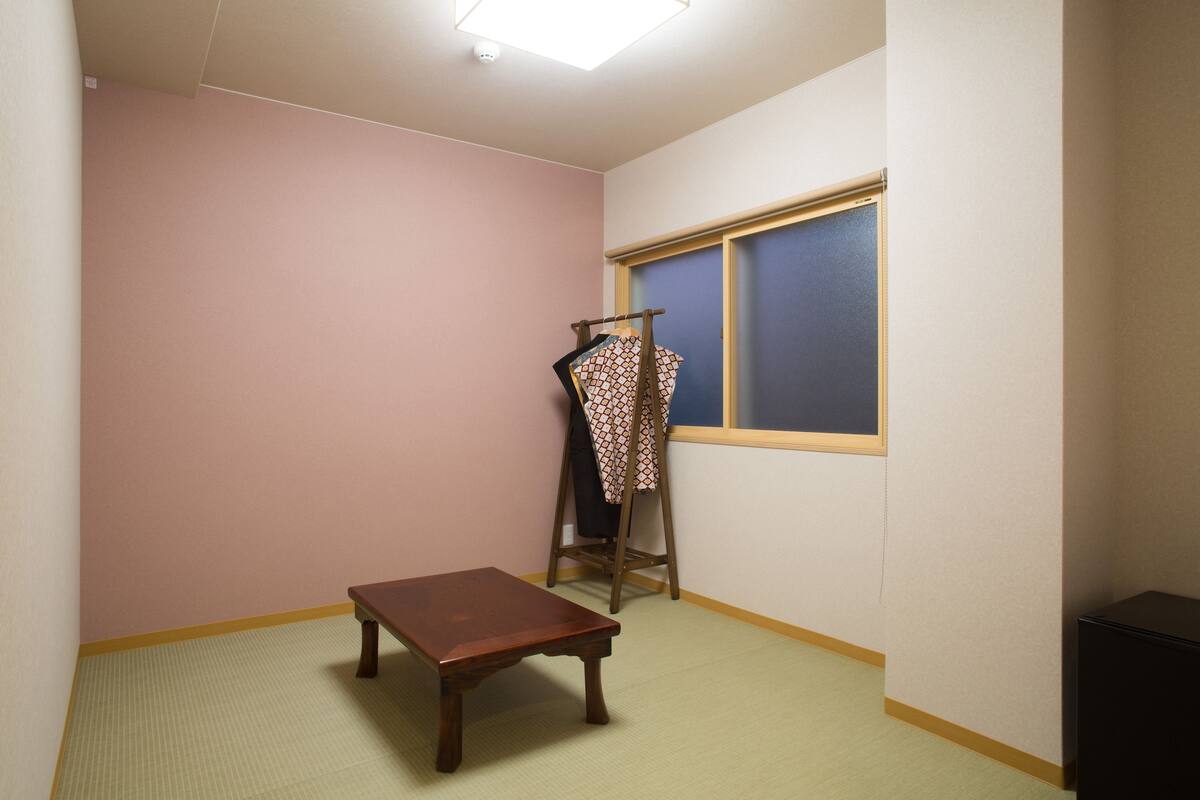 A room with a wooden bench, a clothes rack with patterned items, a window with blinds, and a light fixture on the ceiling.