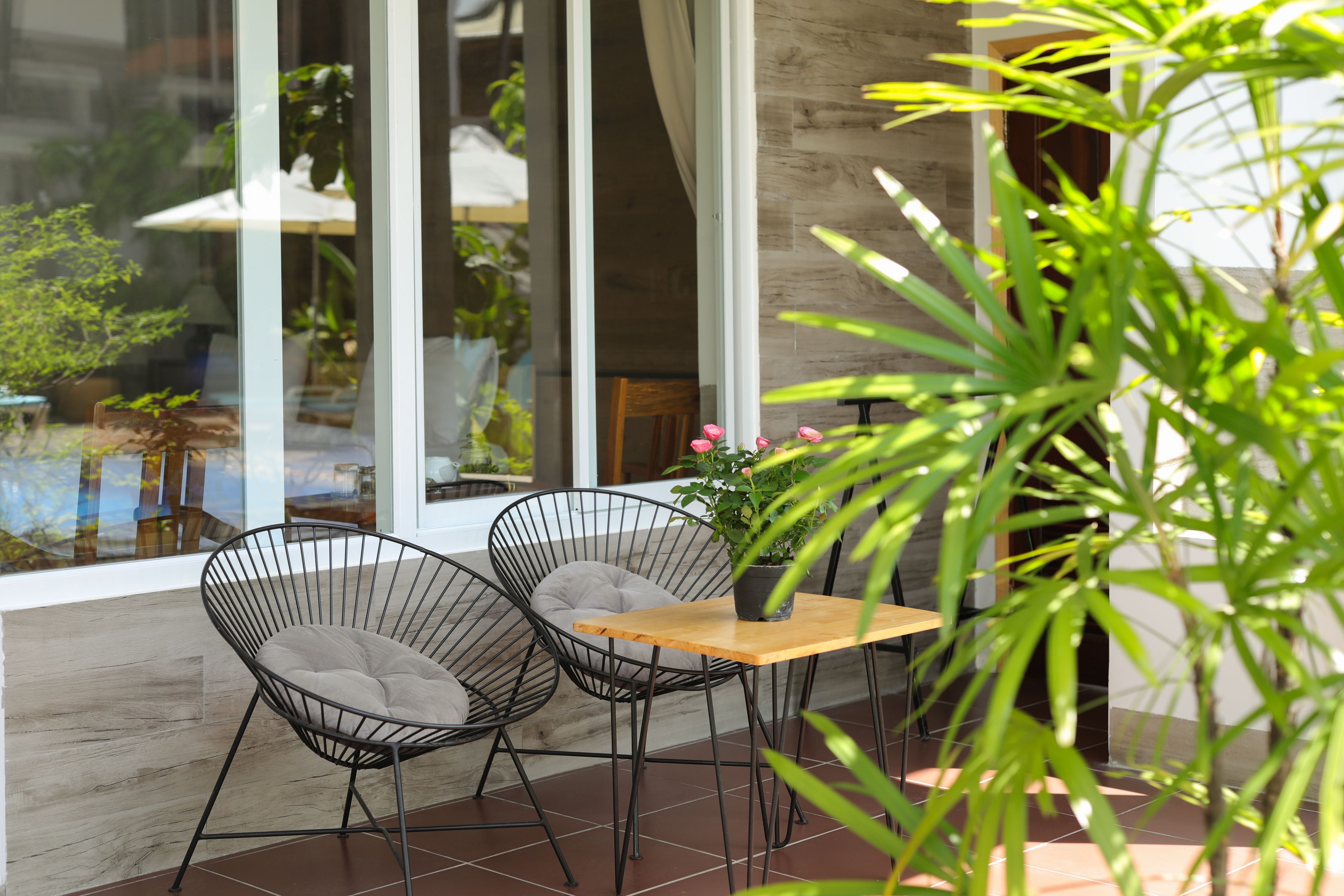 A modern outdoor seating area with two wireframe chairs, a wooden table, and a potted plant with pink flowers.