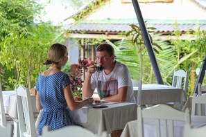 A dining table with a white tablecloth, surrounded by white chairs, in a tropical setting with lush greenery and a traditional tiled roof structure in the background.