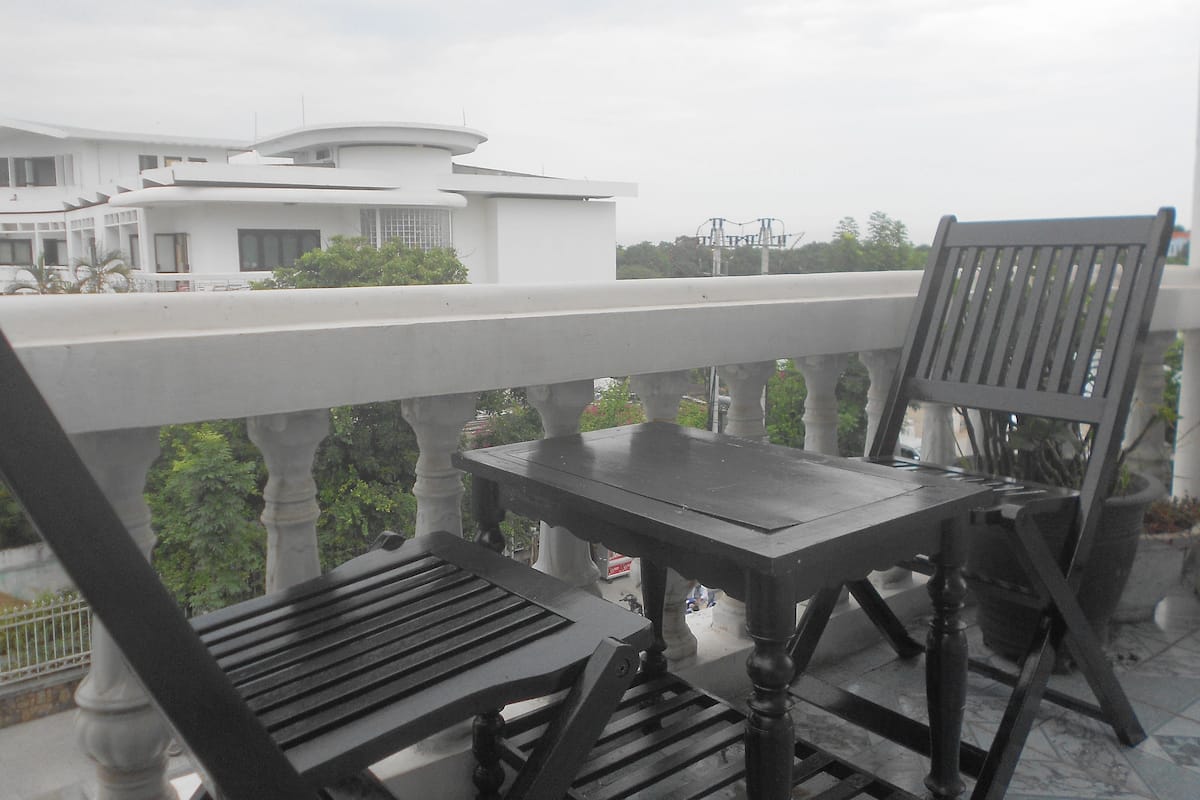 A balcony with a table and two chairs, overlooking a white building and greenery.