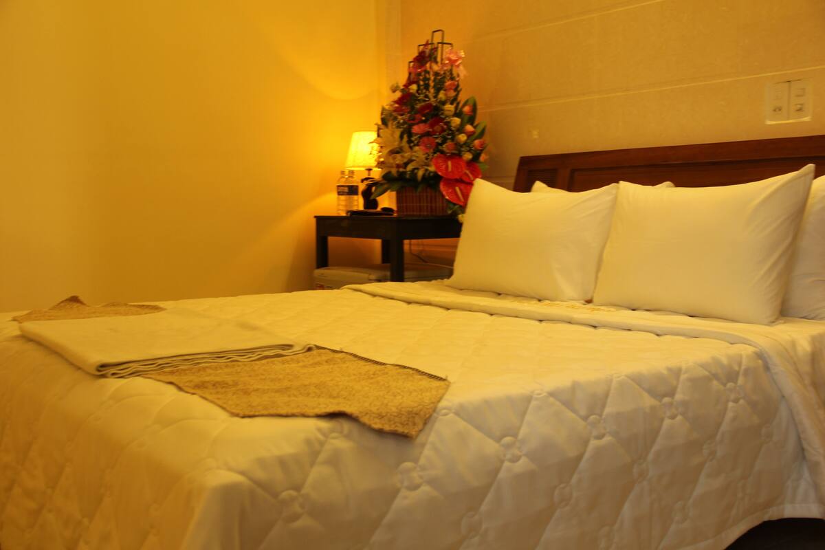 A neatly made bed with white linens and pillows, a bedside table with a lamp and a floral arrangement, and a wall-mounted light switch.