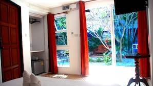 A hotel room with a bed, a small table, a TV, and a sliding glass door leading to a garden. - Ganesha House (Chiang Mai)