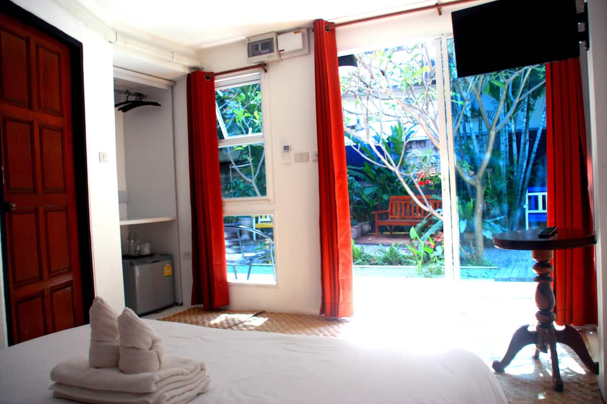 A hotel room with a bed, a small table, a TV, and a sliding glass door leading to a garden.