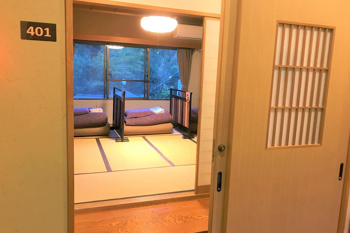 Una habitación japonesa tradicional con suelo de tatami, un futón, una puerta corredera y una ventana con vistas a árboles.