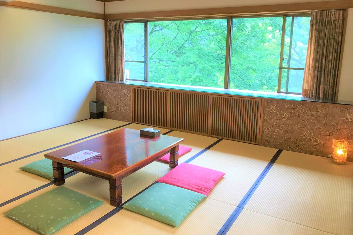 Una habitación de tatami con una mesa baja, cojines y vistas a zonas verdes por una ventana.