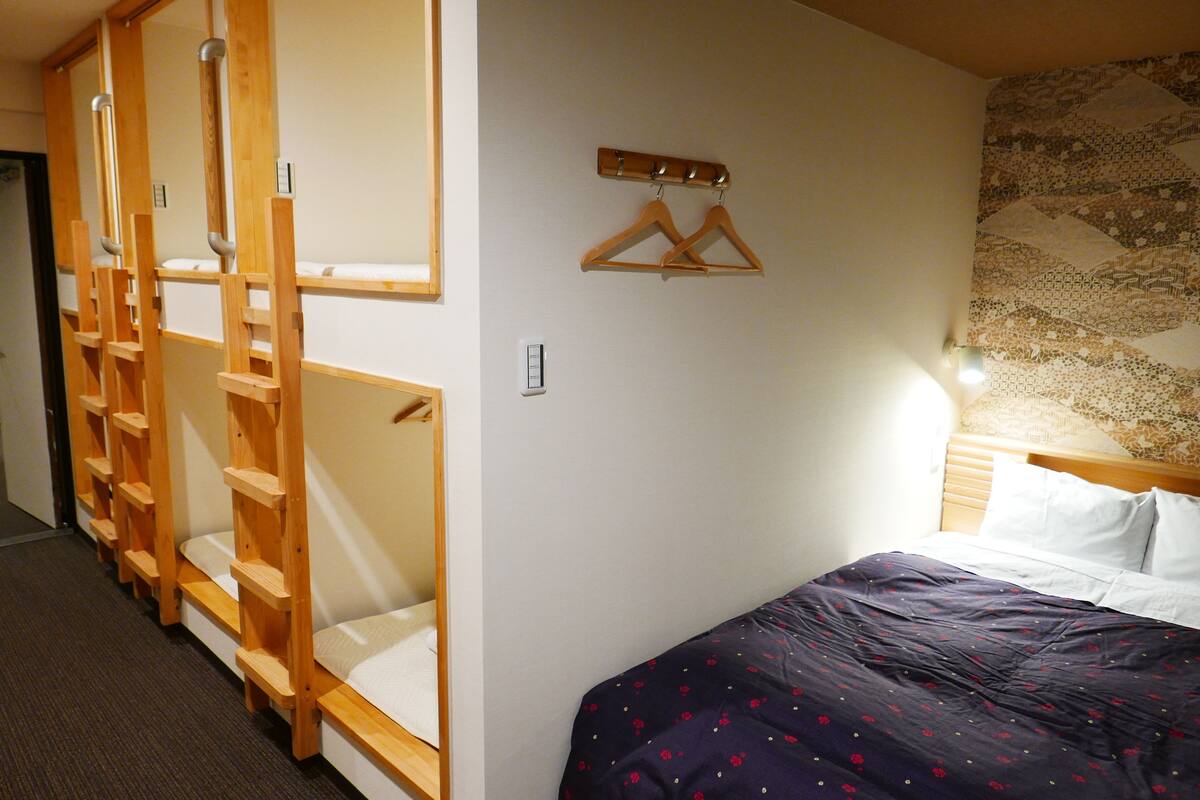 Una habitación con literas, una cama con una colcha estampada, perchas de madera en la pared y un papel tapiz con diseño detrás de la cama.