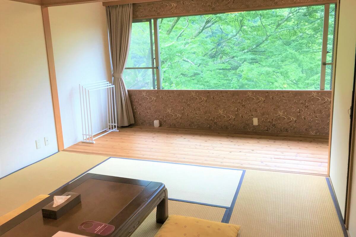 Una habitación japonesa tradicional con suelo de tatami, una mesa baja, un cojín y una ventana con vistas a vegetación.