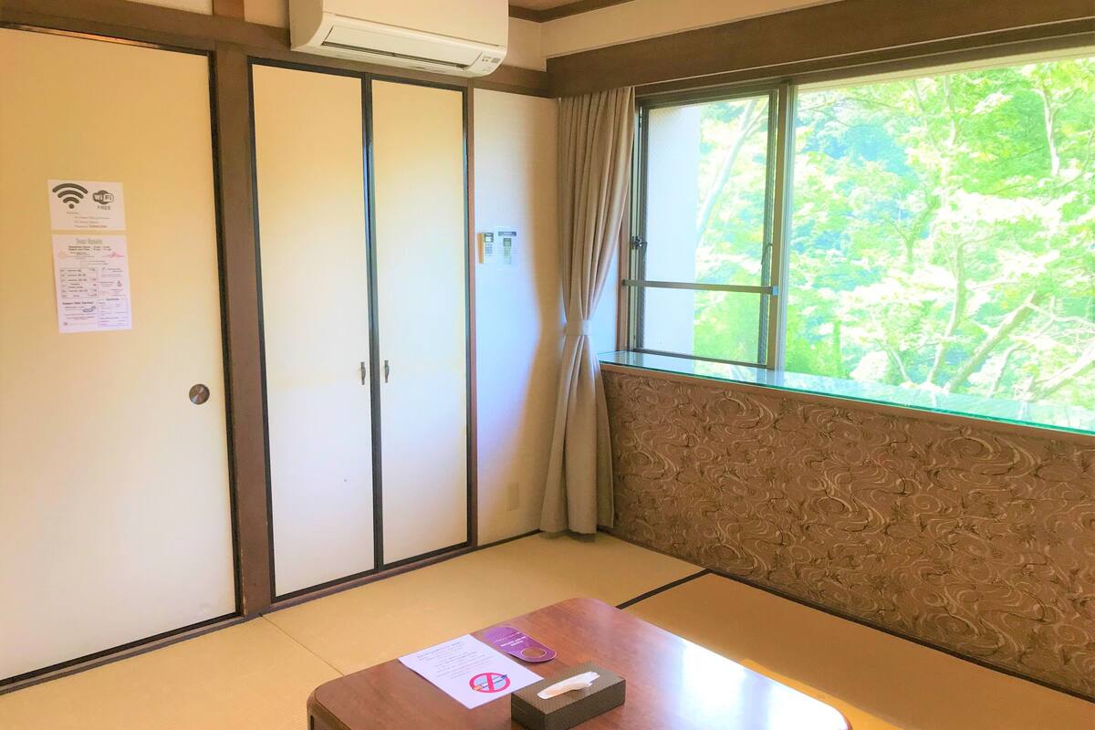 Una habitación con puerta corredera, ventana con vista a follaje, una mesa con una caja de pañuelos y un papel, y aire acondicionado instalado en la pared.