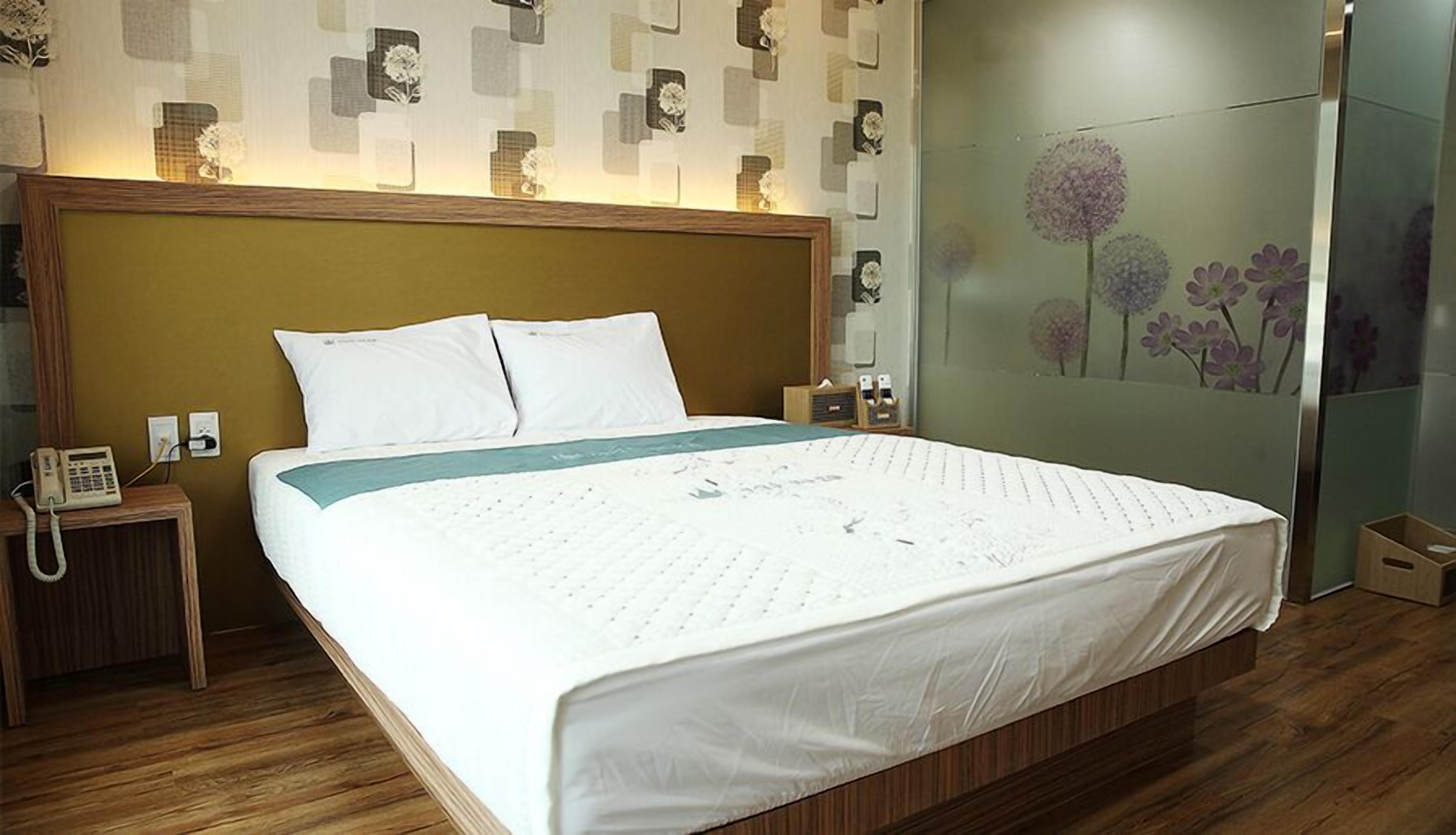Una cama bien hecha con sábanas blancas, un cabecero de madera y una mesita de noche con un teléfono.