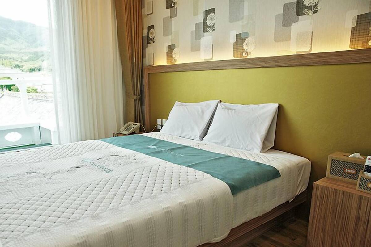 A hotel room with a bed, a bedside table, a telephone, and a window with a view of buildings and hills.