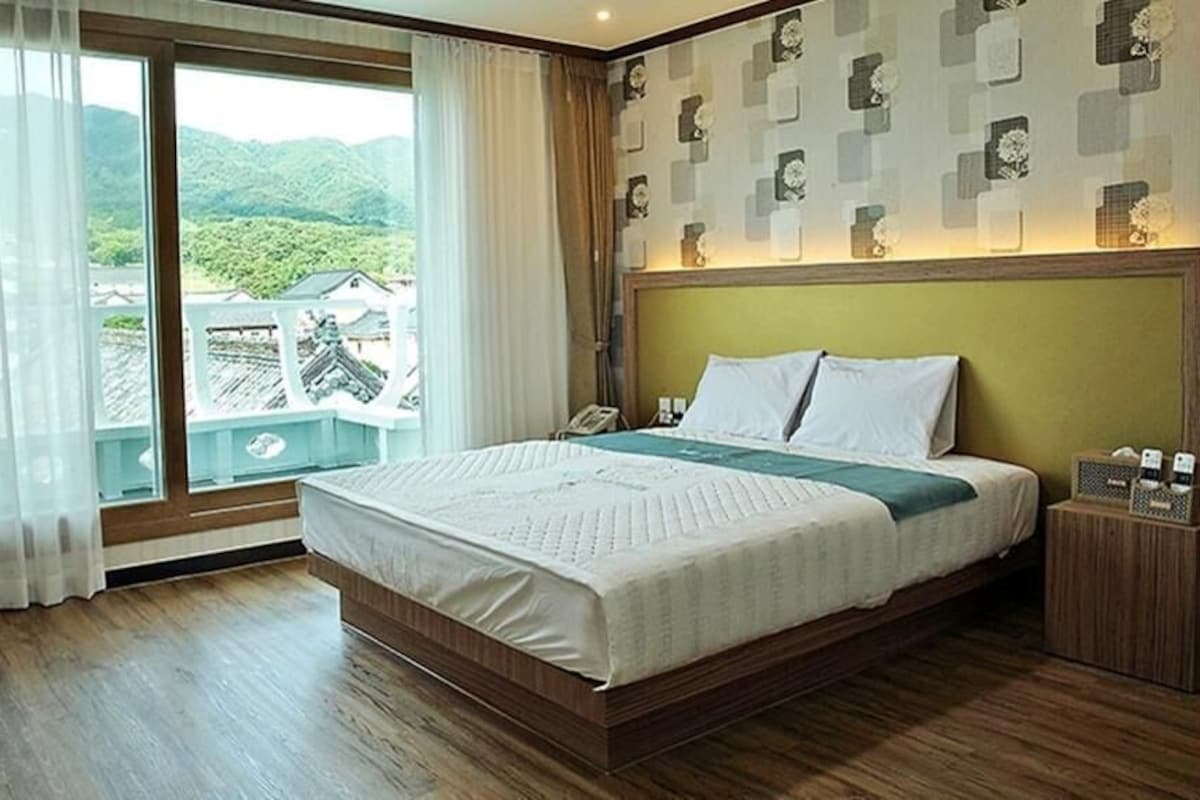 Ein Hotelzimmer mit einem großen Bett, einem Nachttisch, Vorhängen an einem Fenster und Blick auf Grünflächen.