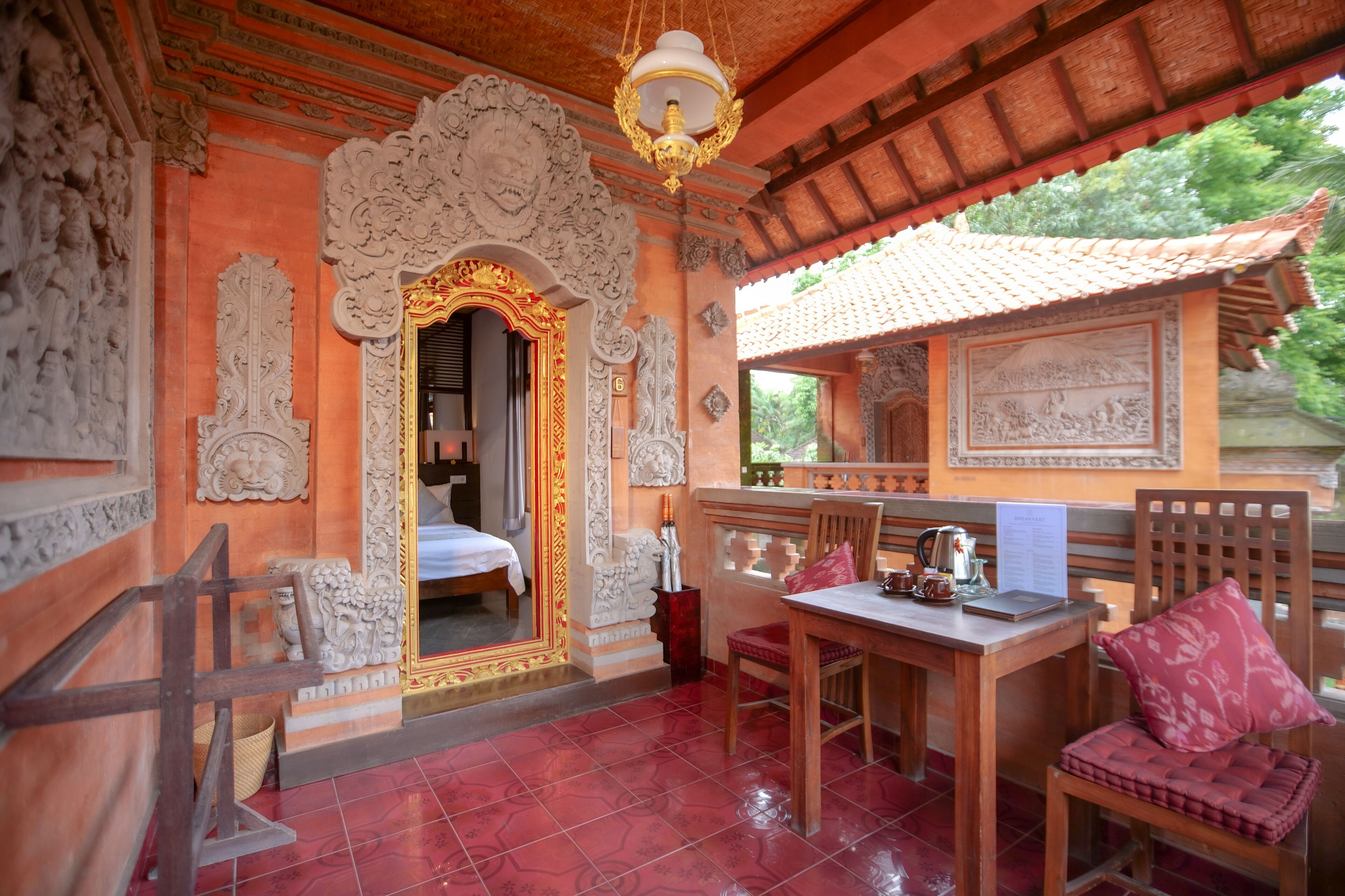 A traditional room with ornate carvings, a bed, a table with a kettle, and a view of greenery outside.