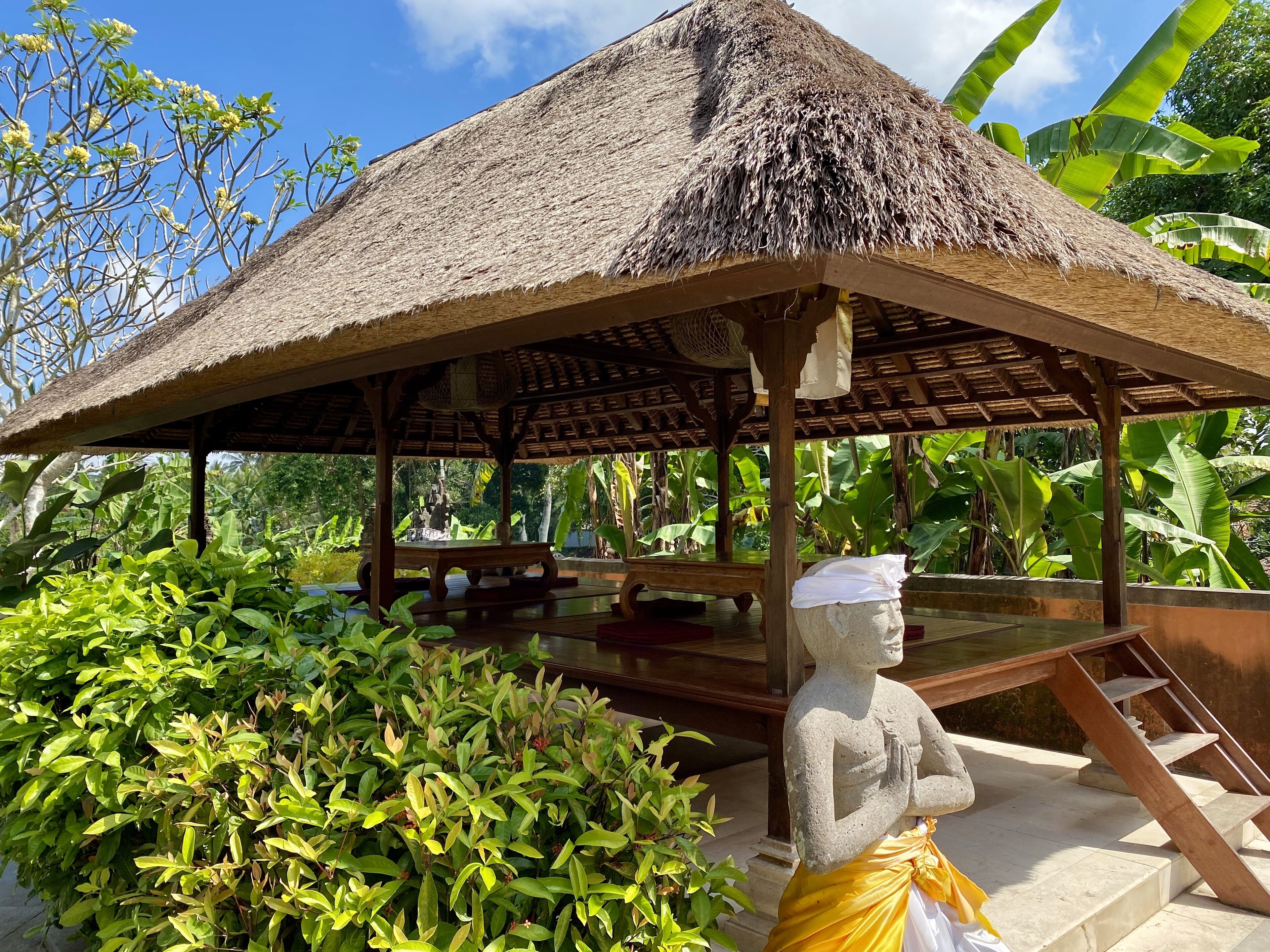 A thatched-roof pavilion with a statue of a figure in traditional attire, surrounded by lush greenery.
