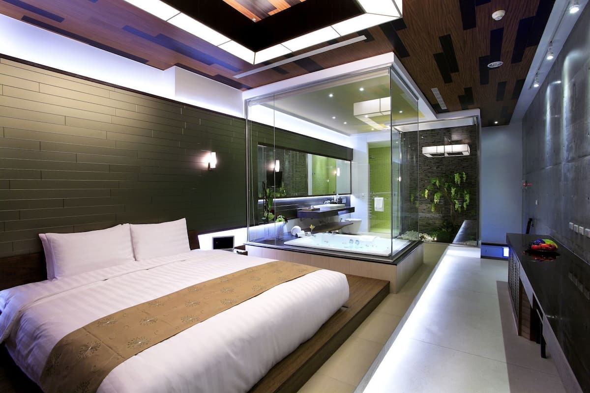 A modern hotel room with a large bed, a glass-enclosed bathroom, and a view of the greenery outside.