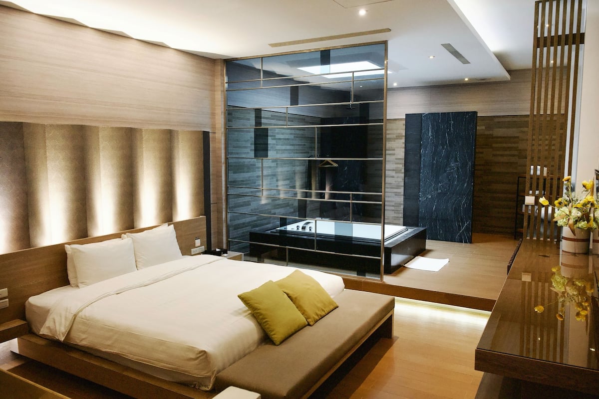A modern bedroom with a large bed, a glass-enclosed bathroom, and a wooden reception desk.