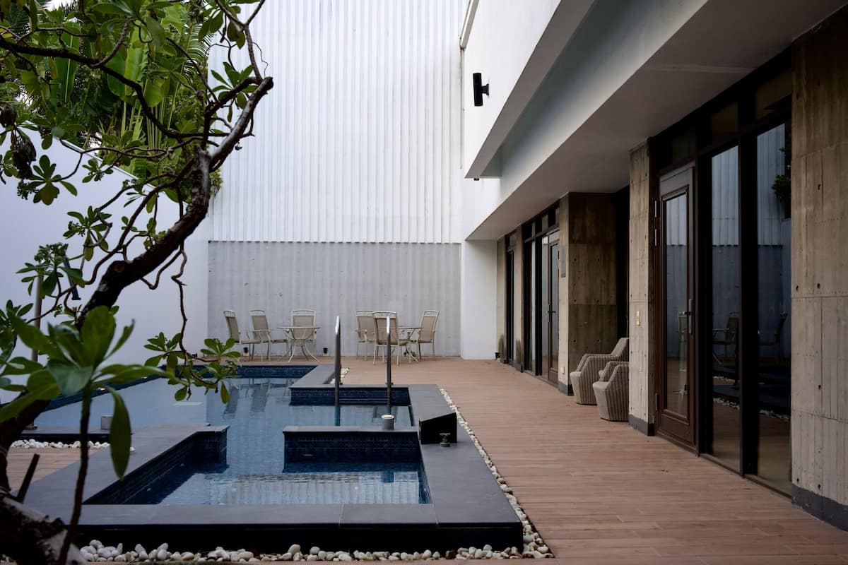 A modern outdoor pool area with a wooden deck, lounge chairs, and a large building in the background.