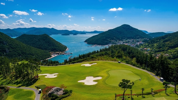 Campo de golf con búnkeres, una masa de agua y un pueblo al fondo.