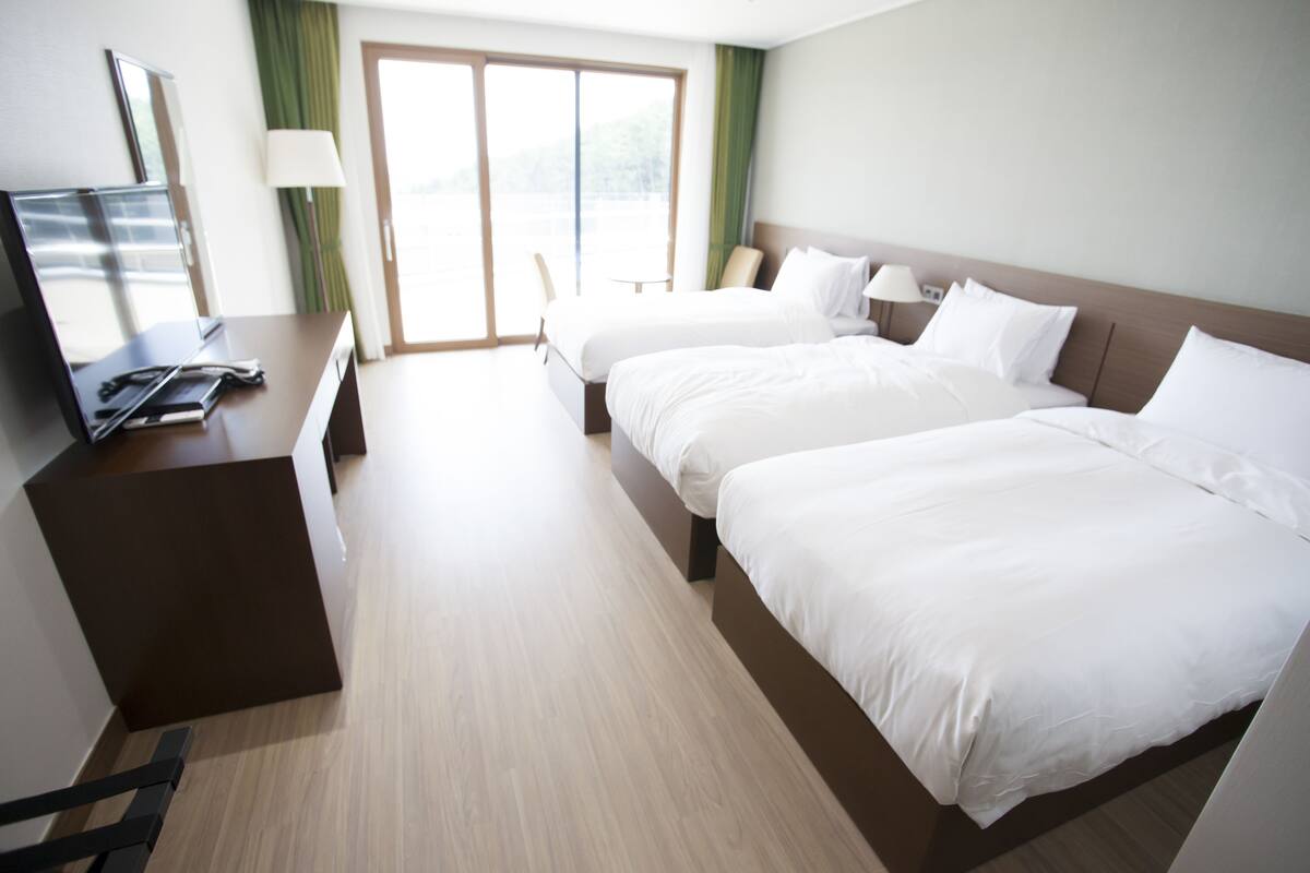 A hotel room with two beds, a desk with a laptop, and a view of the outdoors through a large window.