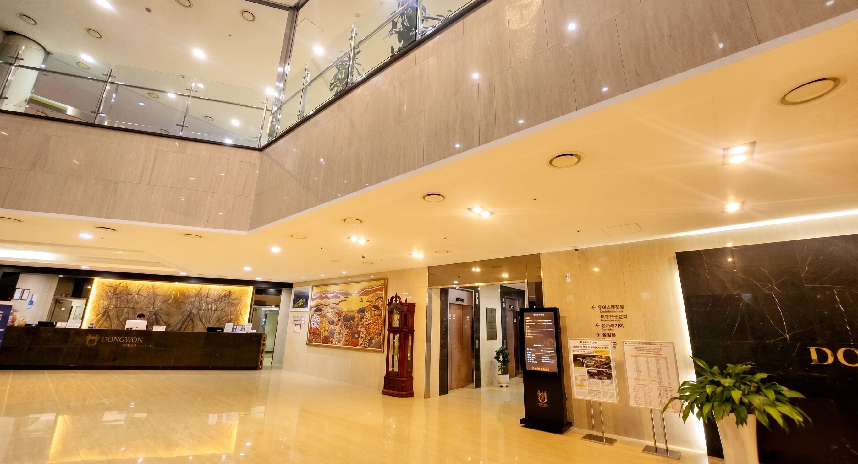 A hotel reception area with a reception desk, a large painting, a clock, and a digital information board.