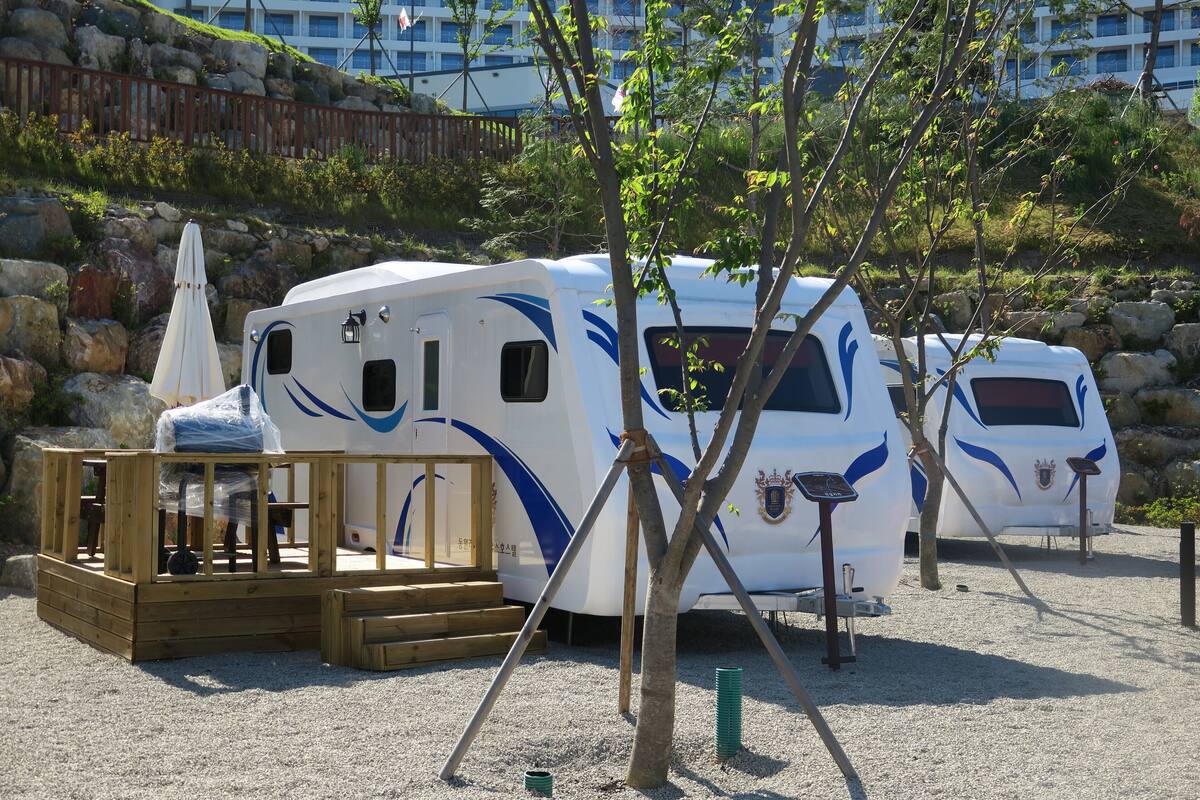 A recreational vehicle (RV) with a wooden deck and a covered area, parked on a gravel surface with trees and a rocky hillside in the background.