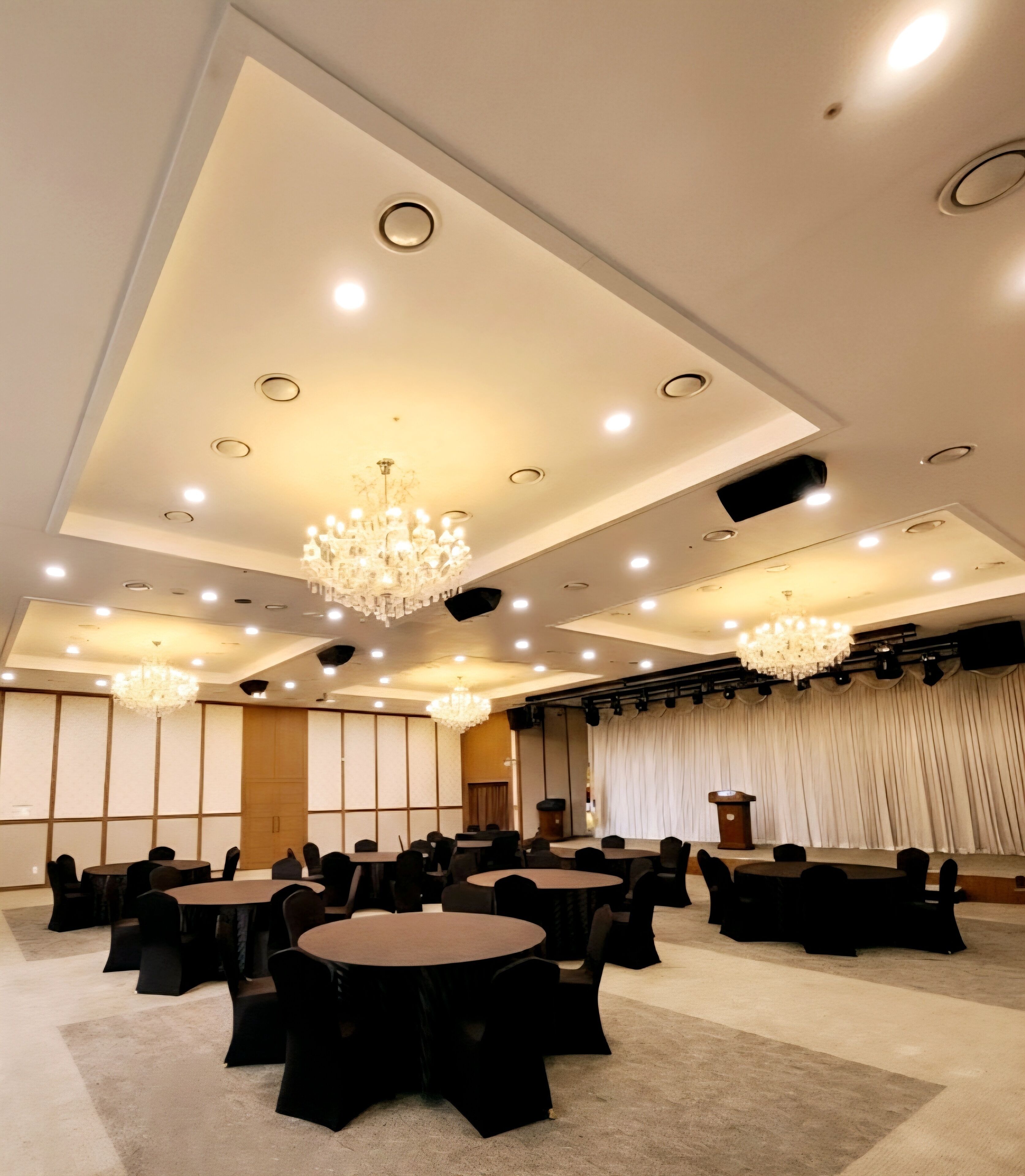 A conference room with round tables, black chairs, chandeliers, and a stage with curtains.
