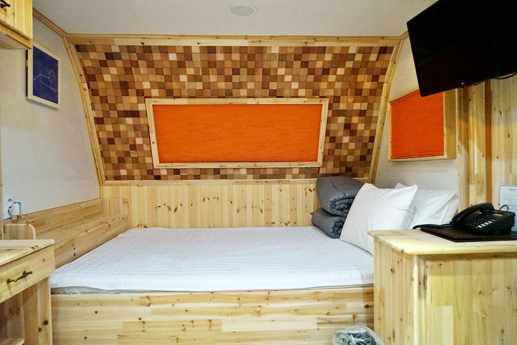 A small, cozy room with a wooden headboard, a bed, a desk with a telephone, and a television mounted on the wall.