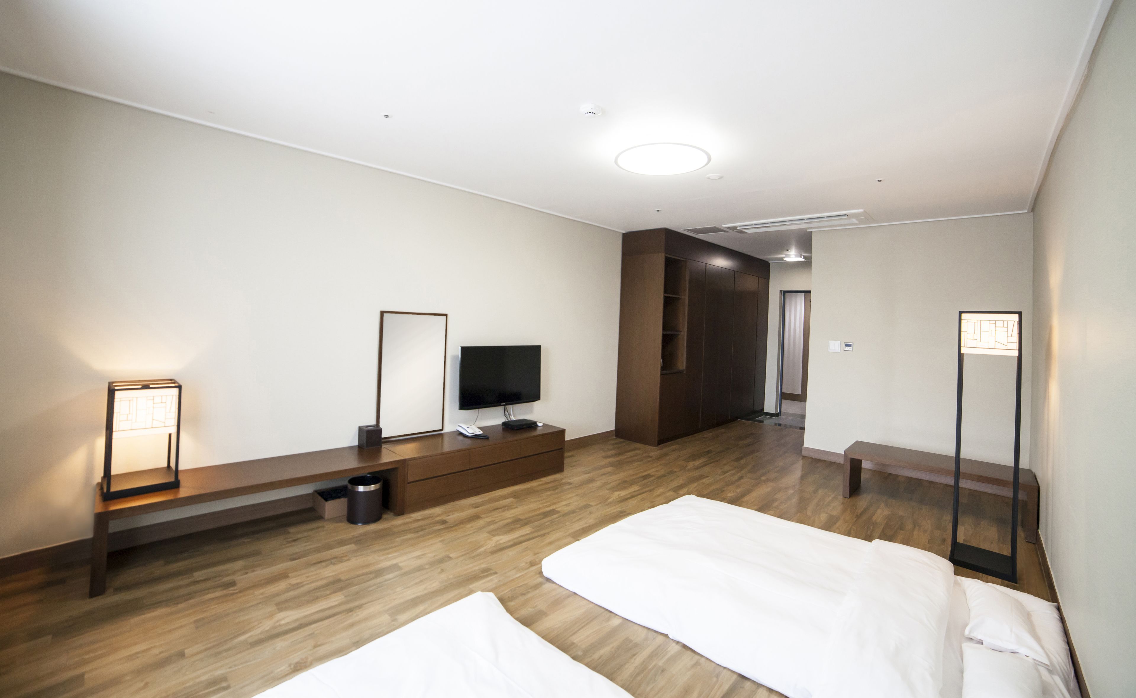 Una habitación de hotel moderna con suelo de madera, televisor de pantalla plana, mesita de noche, una banca y un espejo grande.