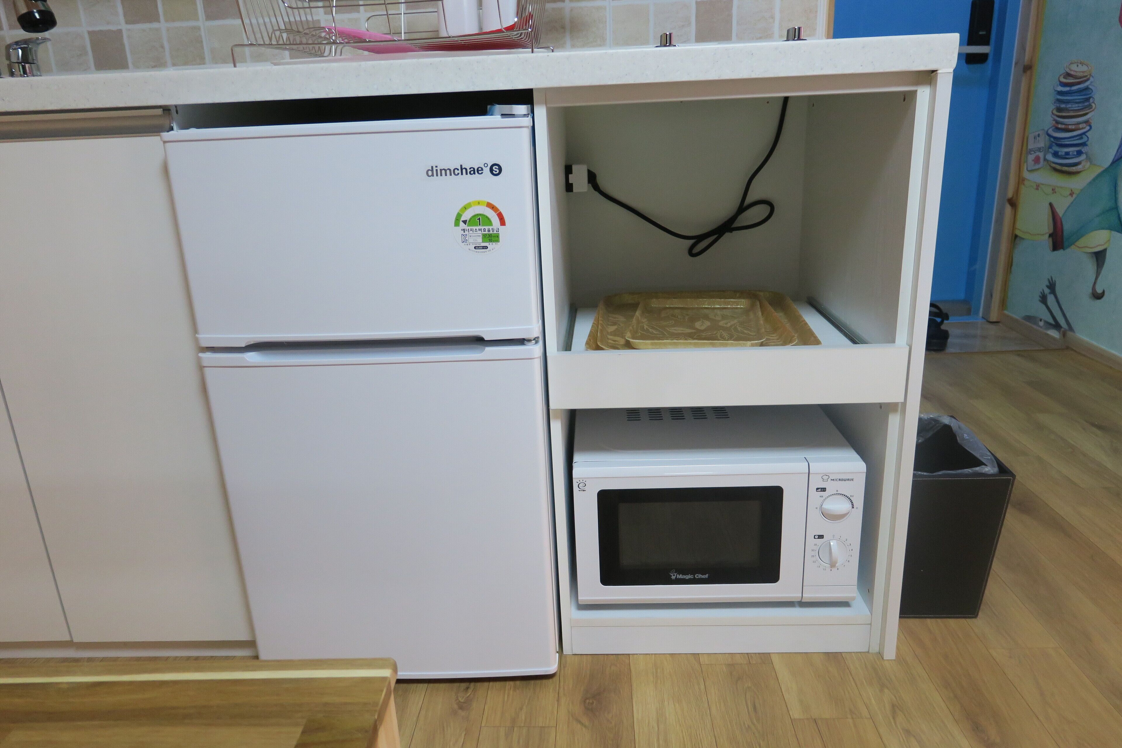 A compact kitchen with a white refrigerator, a microwave, and a shelf with a plastic bag.