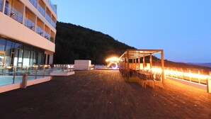 Terraza de madera con mobiliario exterior, un edificio de vidrio y montañas al fondo.
