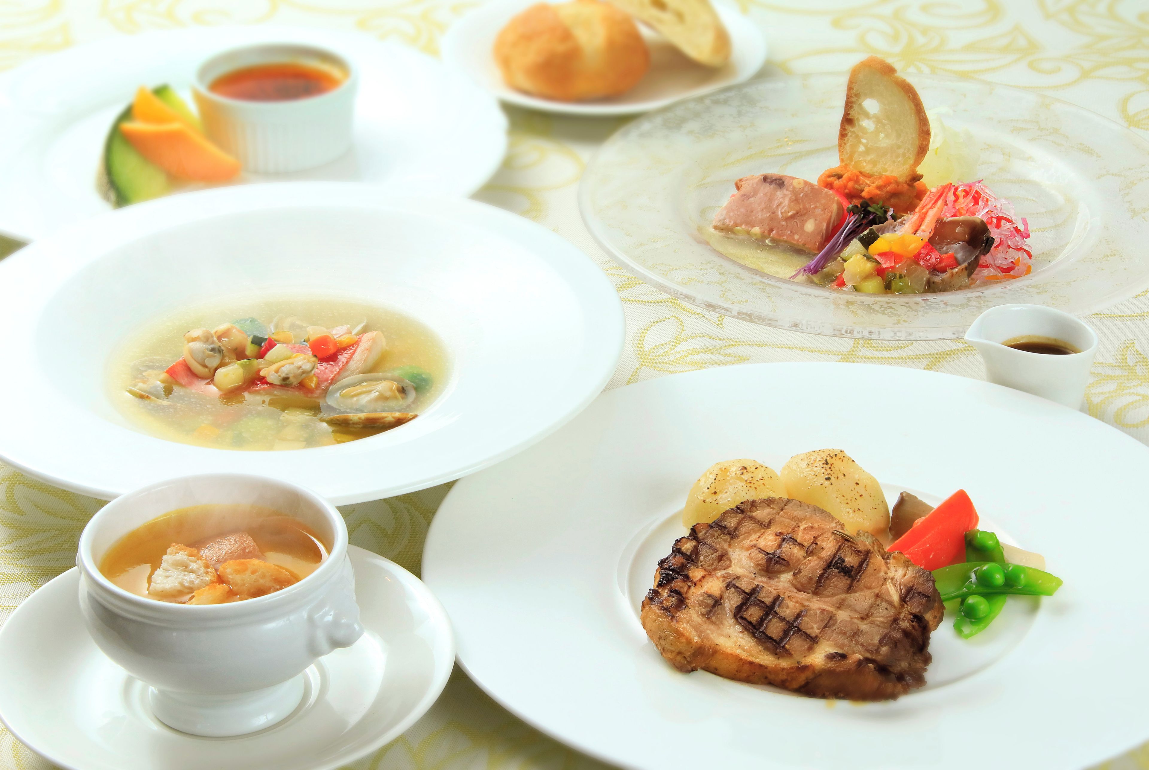 Verschiedene Gerichte, darunter Suppe, gegrilltes Fleisch und Gemüse, auf einem Tisch mit gemustertem Tischläufer.