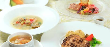 Verschiedene Gerichte, darunter Suppe, gegrilltes Fleisch und Gemüse, auf einem Tisch mit gemustertem Tischläufer.