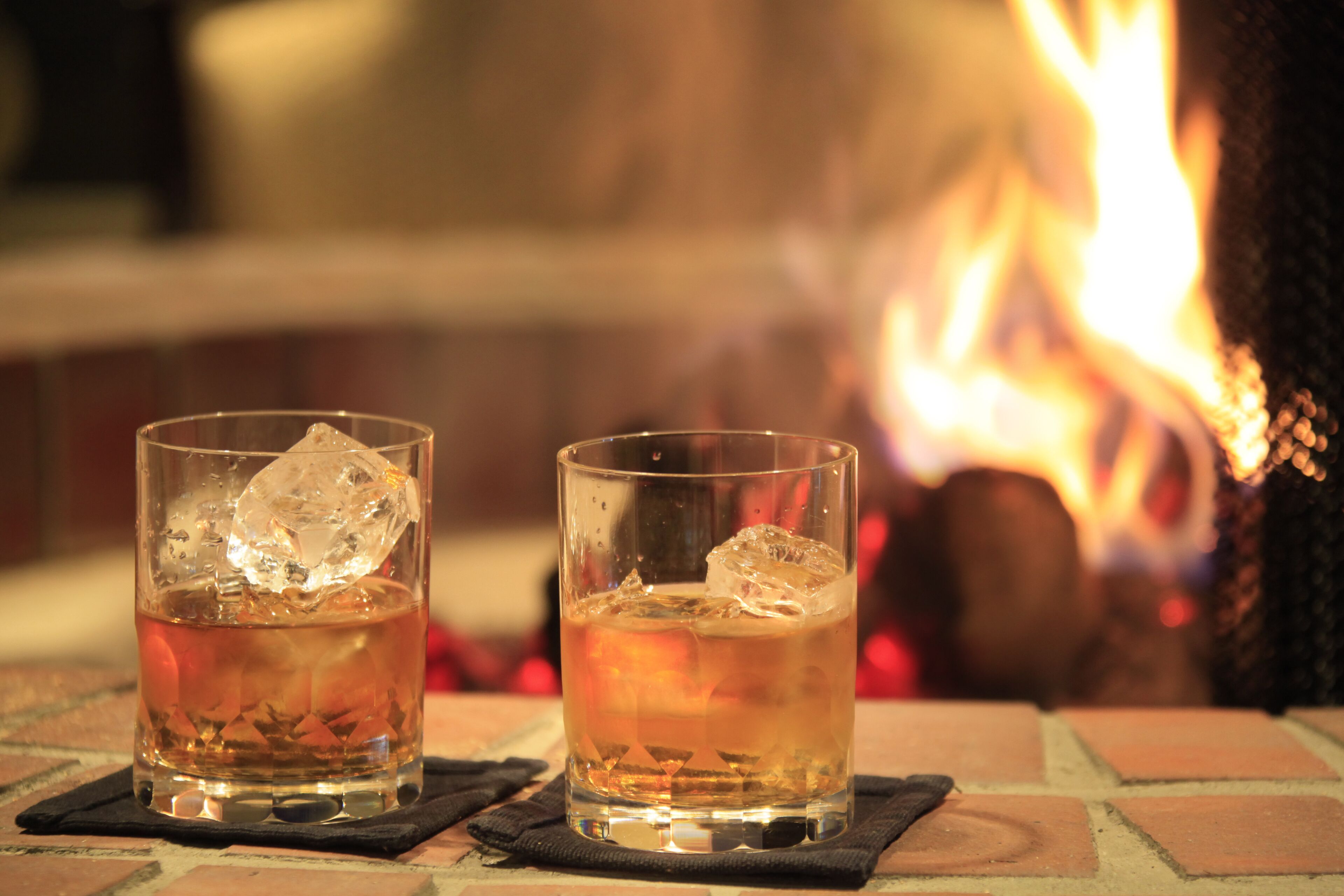 Two glasses of amber-colored beverage with ice cubes on a brick surface, with a blurred background featuring a fire.