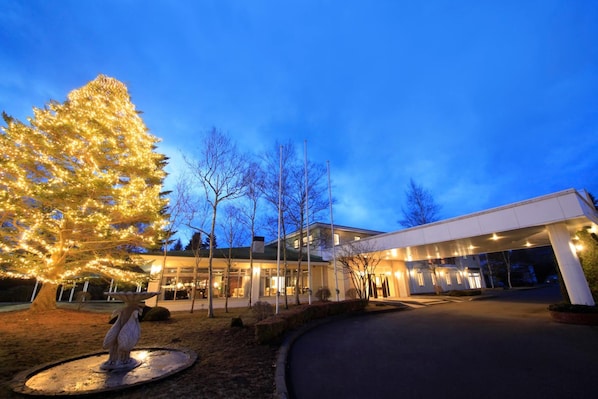 A modern hotel entrance with a well-lit facade, a large illuminated tree, and a statue in the foreground.