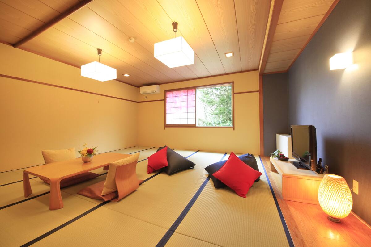 Ett traditionellt japanskt rum med tatamimattor på golvet, ett lågt träbord, röda och svarta kuddar, en tv och en upplyst papperslykta.
