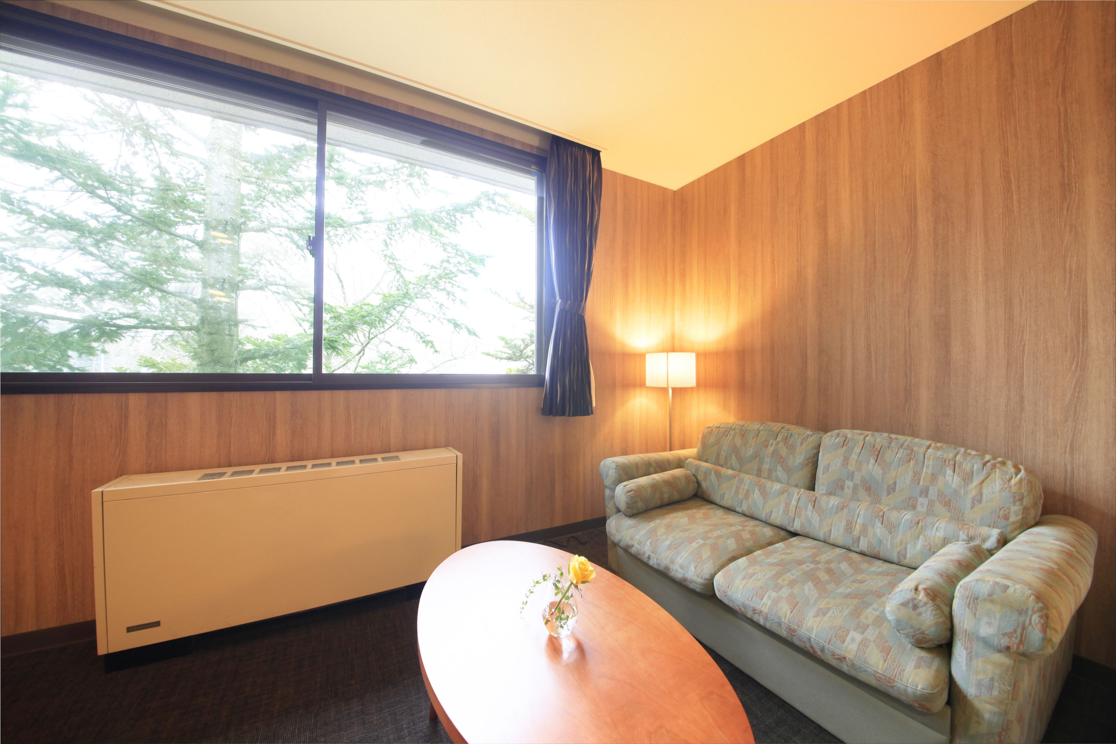 Ein Zimmer mit einer Couch, einem Tisch mit einer Vase, einem Fenster mit Vorhängen und einer Wandlampe.